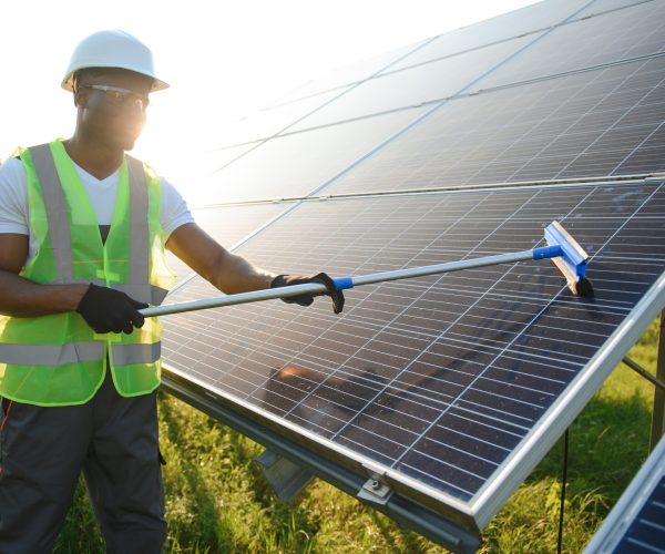 Alternative power plant worker in uniform cleaning solar panels with mop. Handsome African-American taking care of equipment