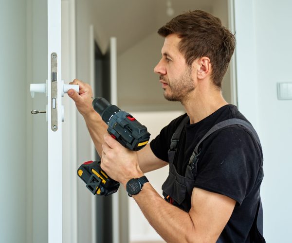 Man holds cordless drill and installs white door handle in modern interior. Handyman fitting door in apartment, using screwdriver. Concept of hardware installation during renovation