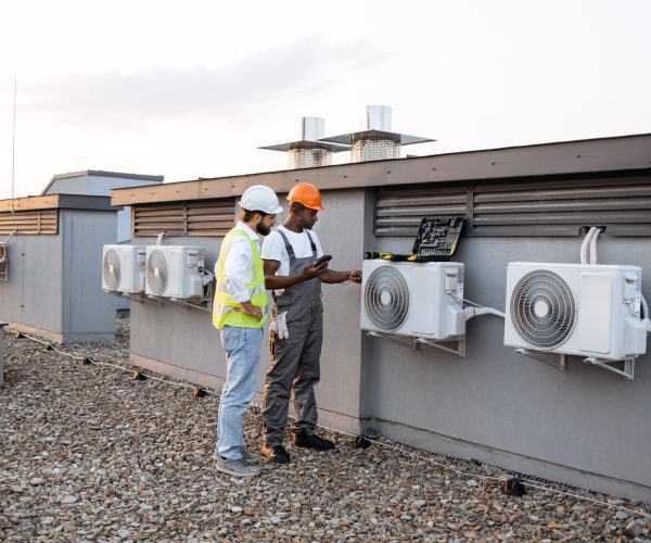 Group of multicultural construction workers on roof inspecting cooling system together using testing tool. Large and gray air conditioning unit with multiple ventilation openings and pipes.